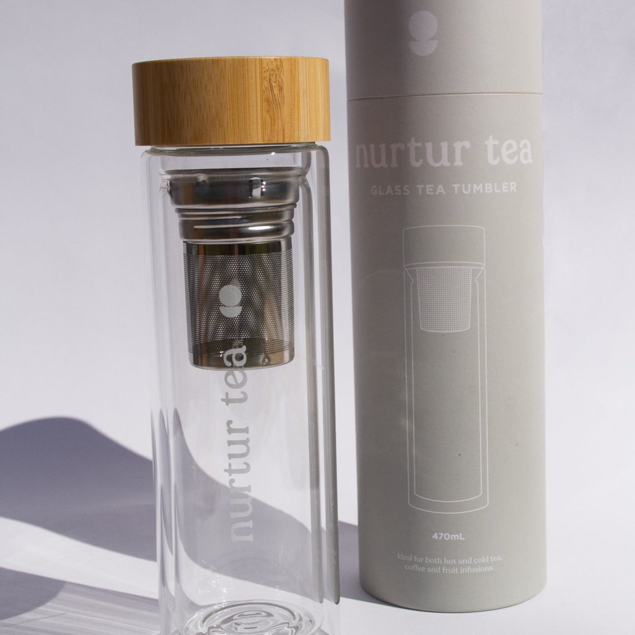 Nurtur tea glass tea infuser with bamboo lid standing upright next to amd slightly in fornt of packaging, grey canister with white writing