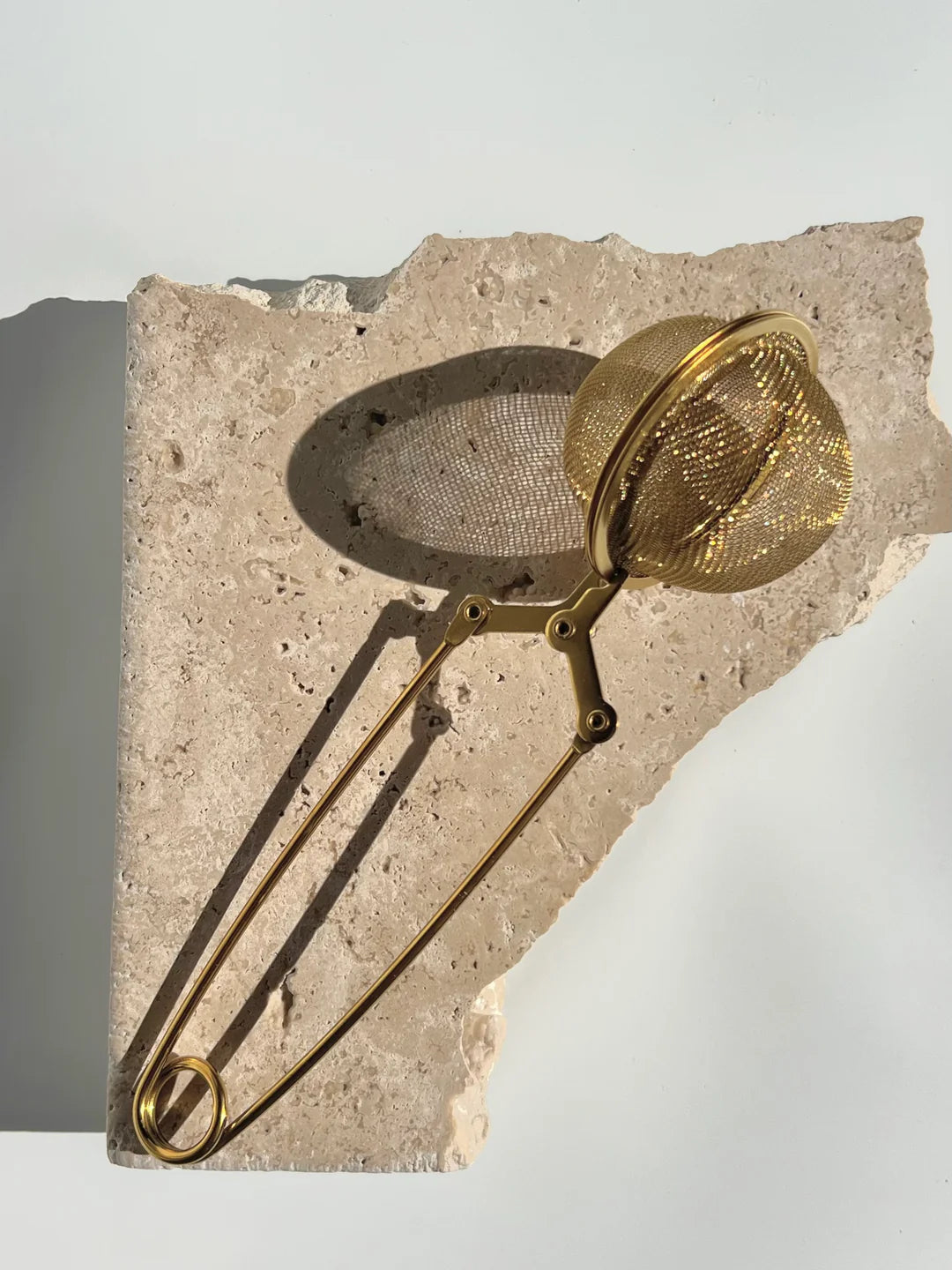 Gold tea strainer with spring handle laying on piece of stone 