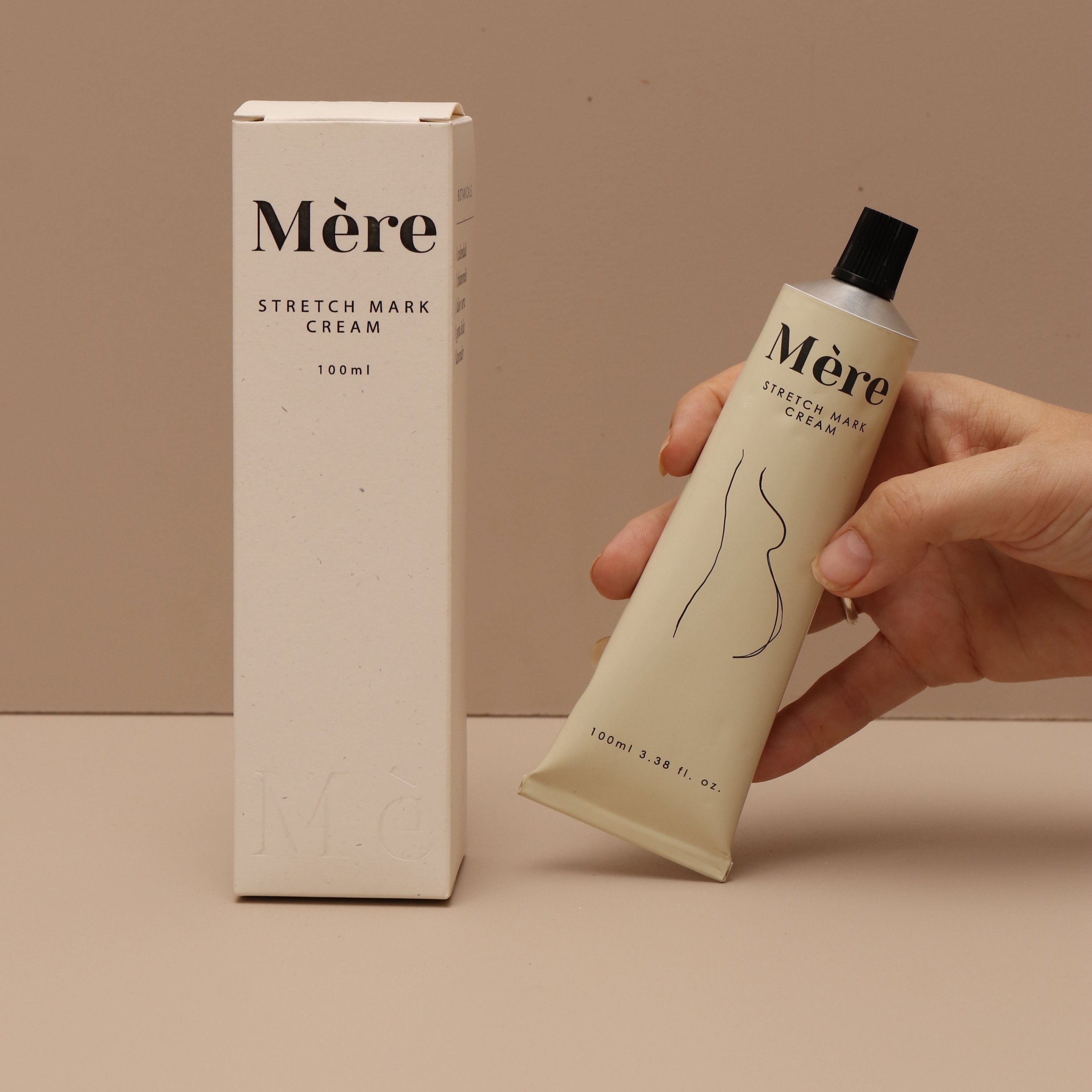 Hand holding tube of mere botanicals stretchmark cream, cream coloured tube with outline drawing of pregnant female body, packing shown next to product, white box with black writing